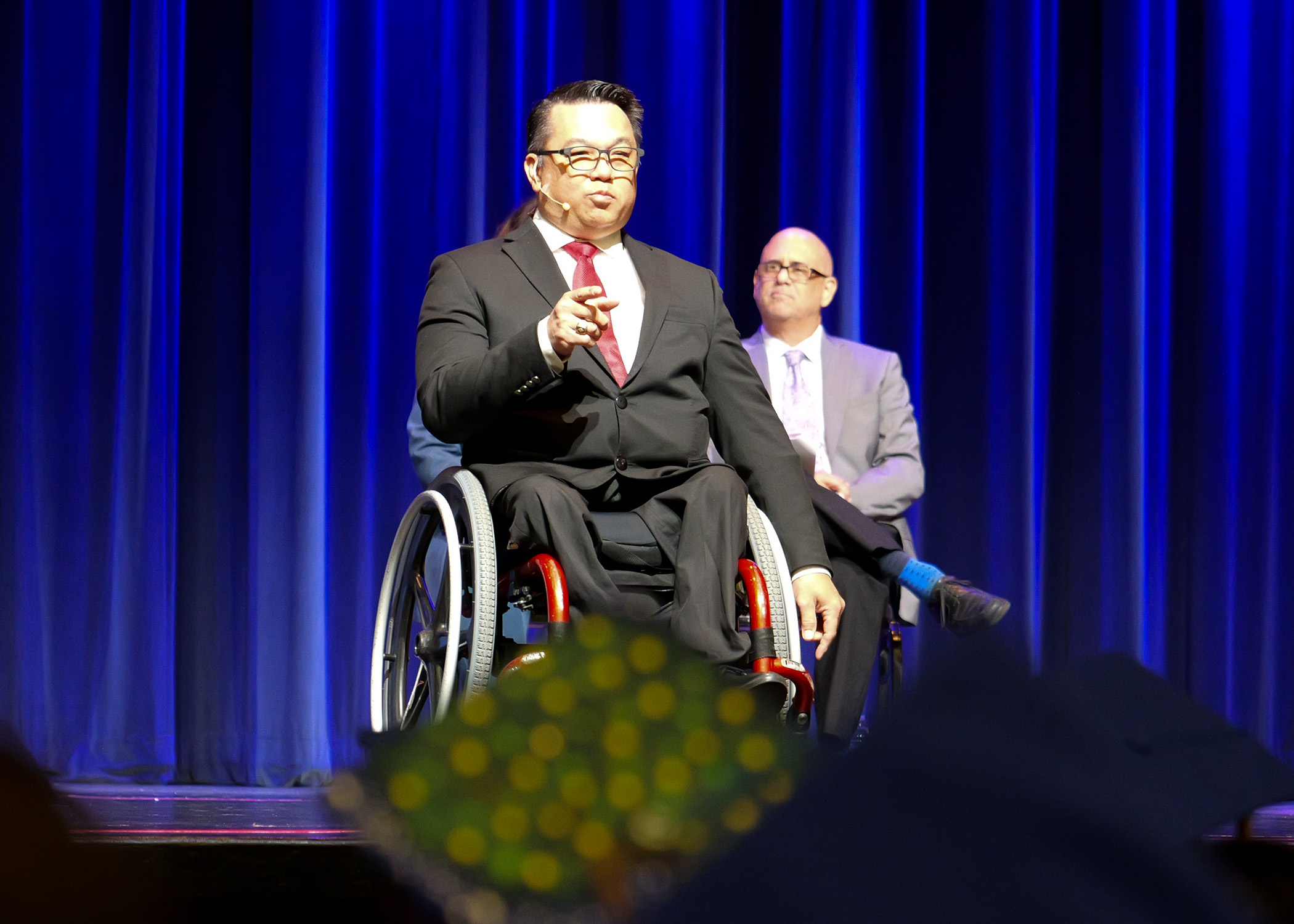 Dr. Stefan Bean gestures while delivering a speech to ACCESS graduates during a commencement ceremony, with decorated caps visible in the foreground.