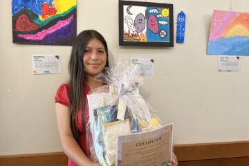 Leah Marie Felix holding a gift basket and certificate in front of her winning artwork — a colorful drawing of lungs promoting a tobacco-free lifestyle — at the 2025 TUPE Art Showcase.