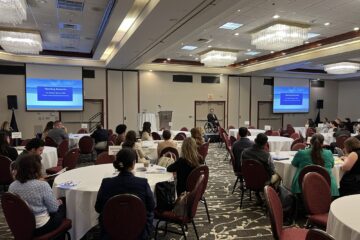 Dr. Stefan Bean speaks on stage during his opening remarks. A presentation slide behind him displays his name and title.
