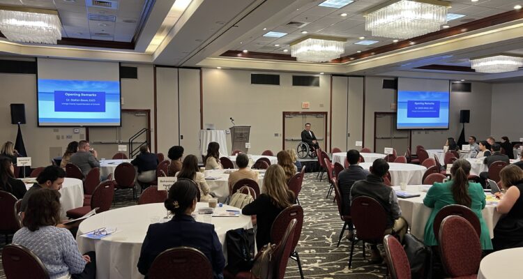 Dr. Stefan Bean speaks on stage during his opening remarks. A presentation slide behind him displays his name and title.